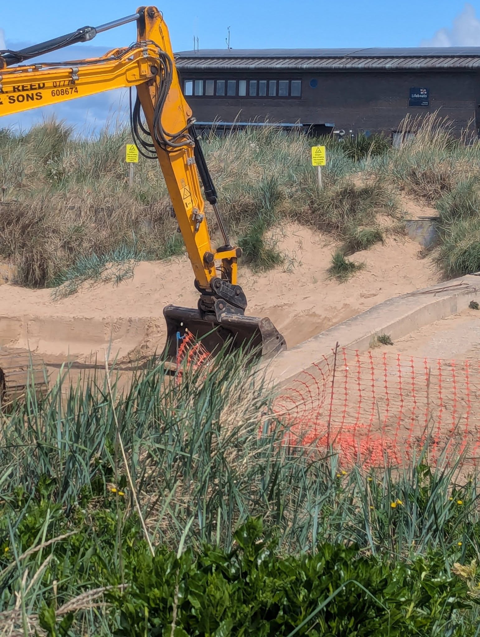 A yellow excavator working on Hoylake beach in front of the RNLI lifeboat station, with orange safety fencing and freshly turned sand