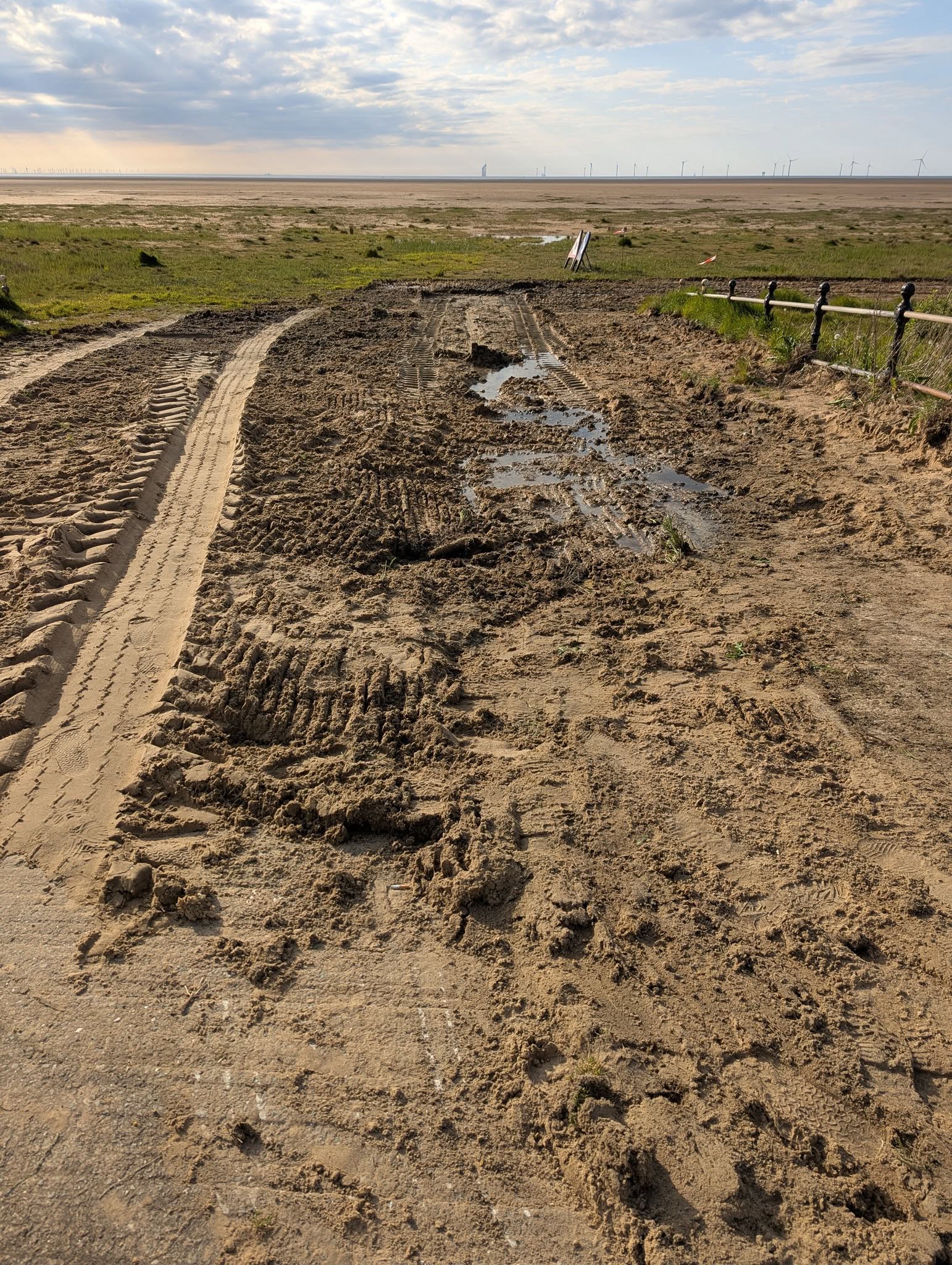 Tractor tyre tracks churned through the cleared saltmarsh at Hoylake, leading towards the Dee Estuary horizon with distant wind turbines
