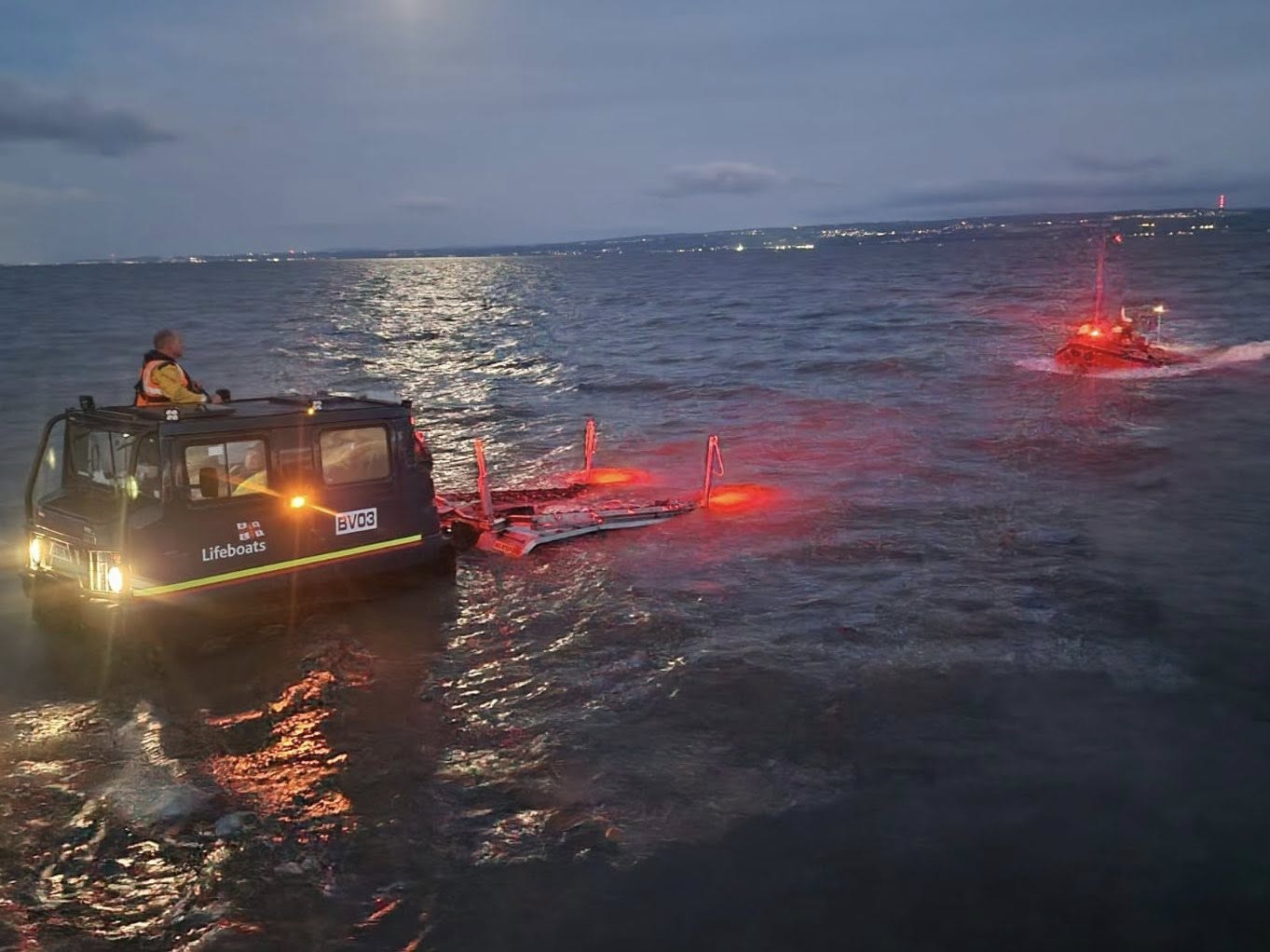 RNLI Hoylake hovercraft launch carriage and an inshore lifeboat with red navigation lights operating in dusk water off the Wirral coast