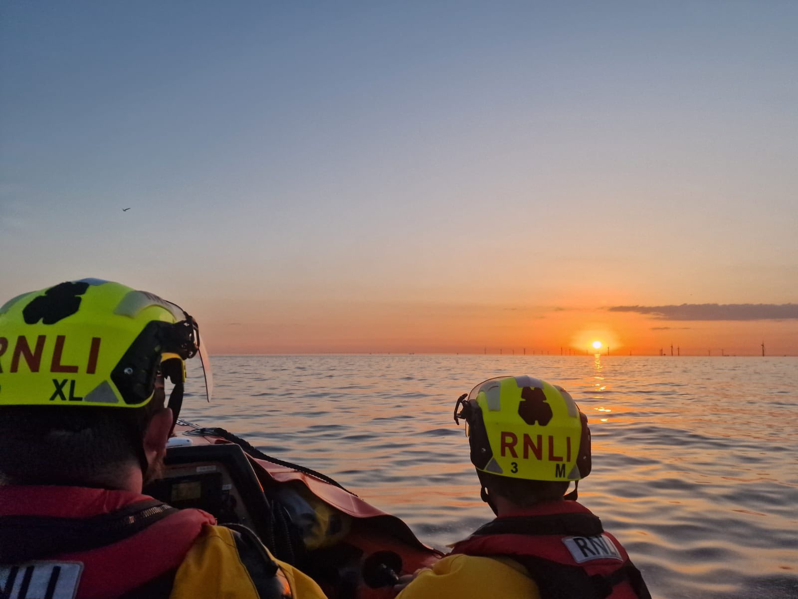 Two RNLI crew in yellow drysuits and yellow helmets on an inshore lifeboat at sunset, looking out across the Irish Sea towards the offshore wind farm