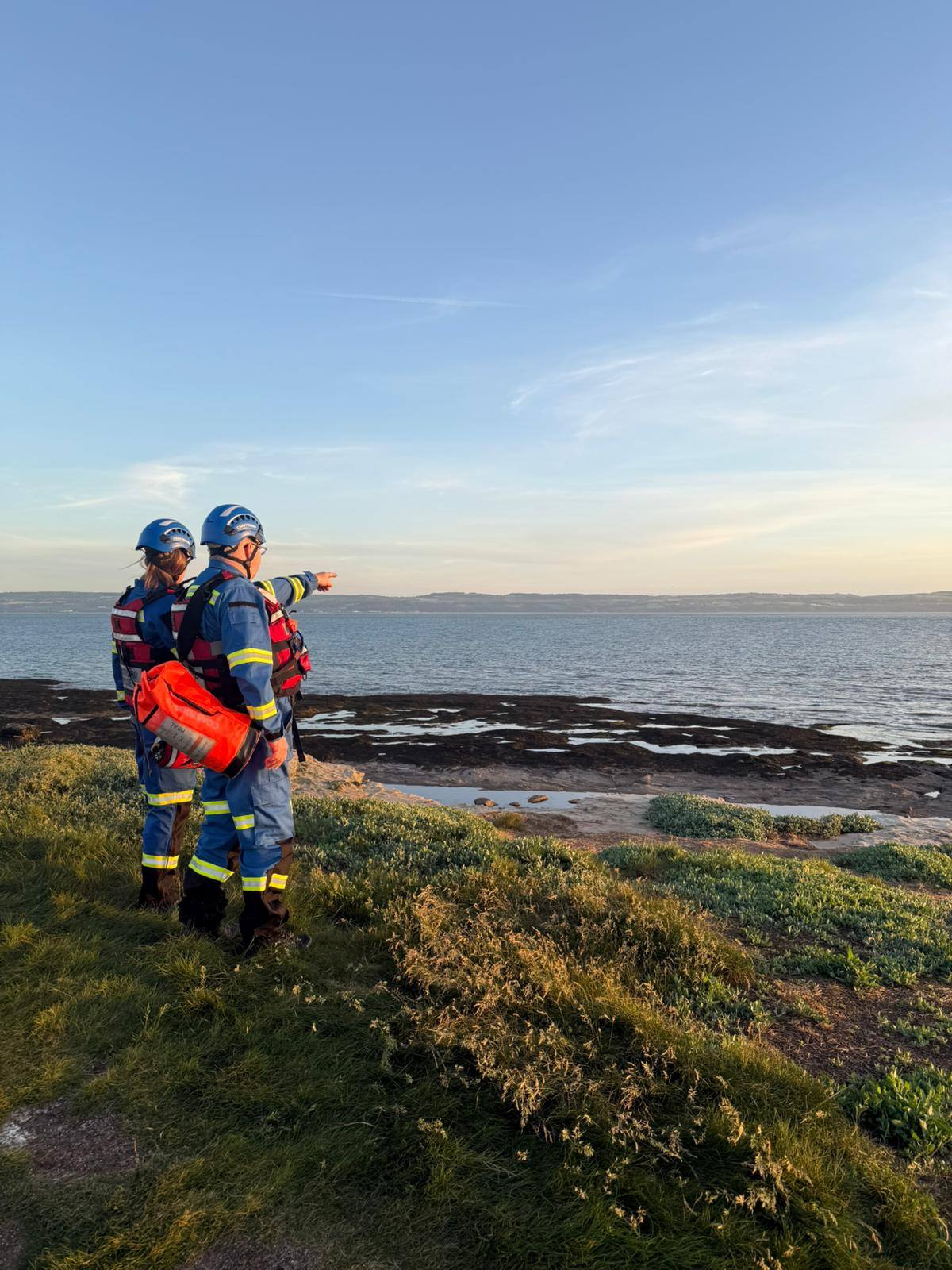 Two Wirral Coastguard Rescue Officers in blue PPE on a grassy clifftop, one pointing out across the Dee Estuary, searching for missing walkers