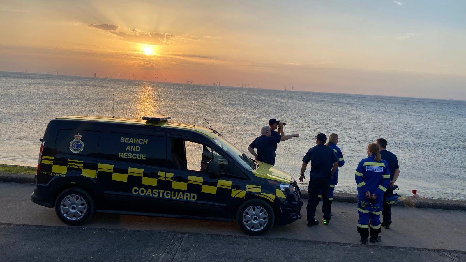 A group of Wirral Coastguard Rescue Officers beside their search and rescue vehicle at sunset, one scanning the sea with binoculars and another pointing out across the water