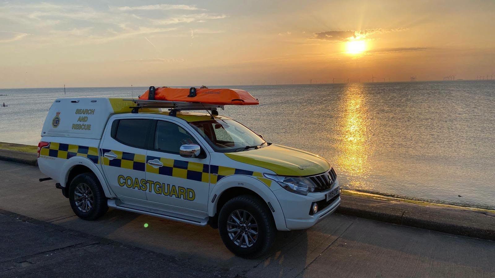 An HM Coastguard search and rescue vehicle with an orange rescue stretcher on its roof, parked on the Wirral promenade at sunset with the offshore wind farm on the horizon